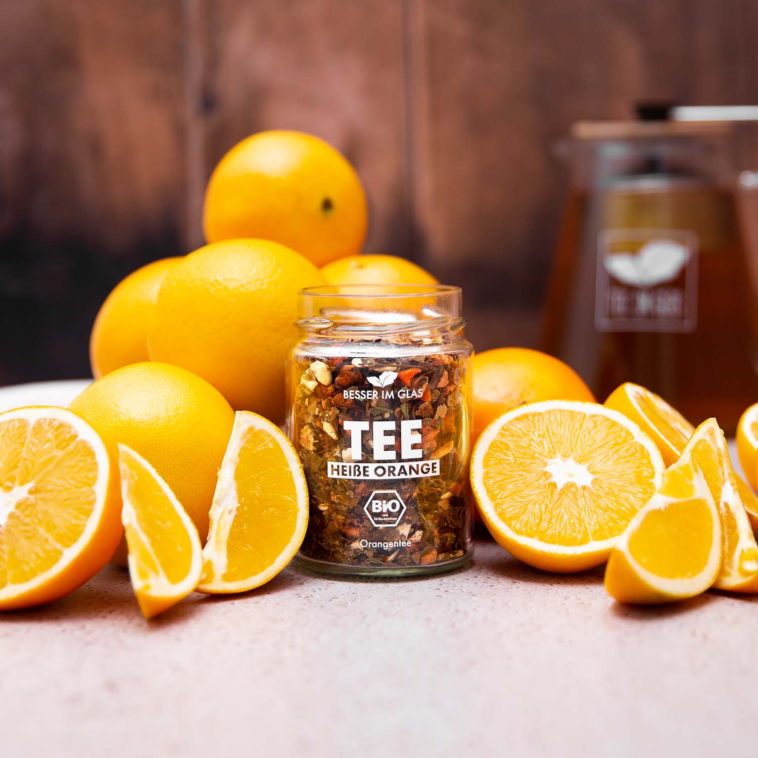 Loser bio Orangen Tee im Glas mit Orangen-Stücken. Bio-Früchte-Kräutertee mit Bio Karotte aromatisiert mit Orangen- und Mandarinengeschmack. Umgeben von Orangen. Im Hintergrund ist eine Teekanne zu sehen. In der Glaskanne befindet sich ein Teesieb mit abnehmbaren Bambus Deckel. Bio Tee ohne Zucker