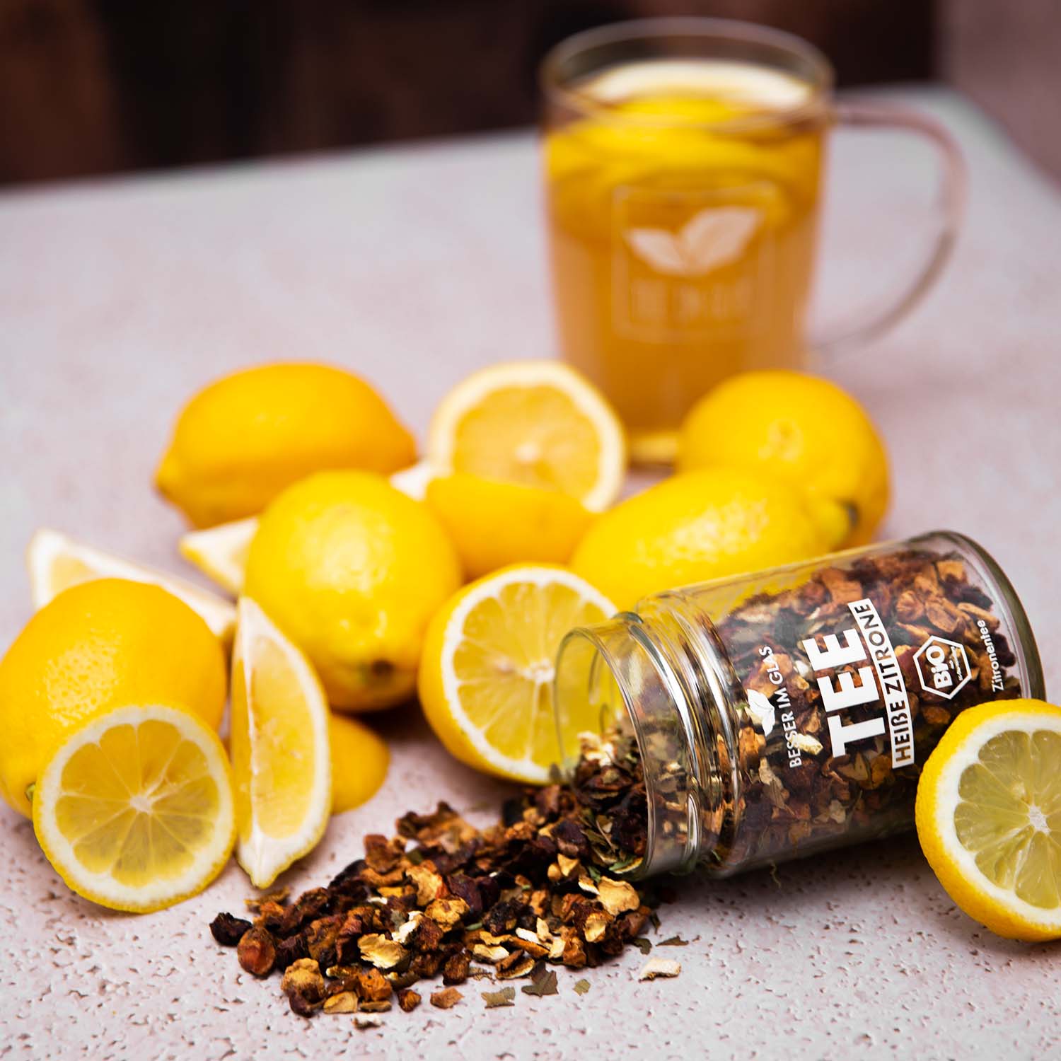 Loser bio Zitronen Tee im Glas mit Zitronen-Stücken. Bio-Früchte-Kräuterteemischung aromatisiert mit Zitronengeschmack. Umgeben von Zitronen. Außerdem ist eine Teetasse zu sehen. In dem Teeglas mit Henkel befinden sich Zitronenscheiben. Bio Tee ohne Zucker.