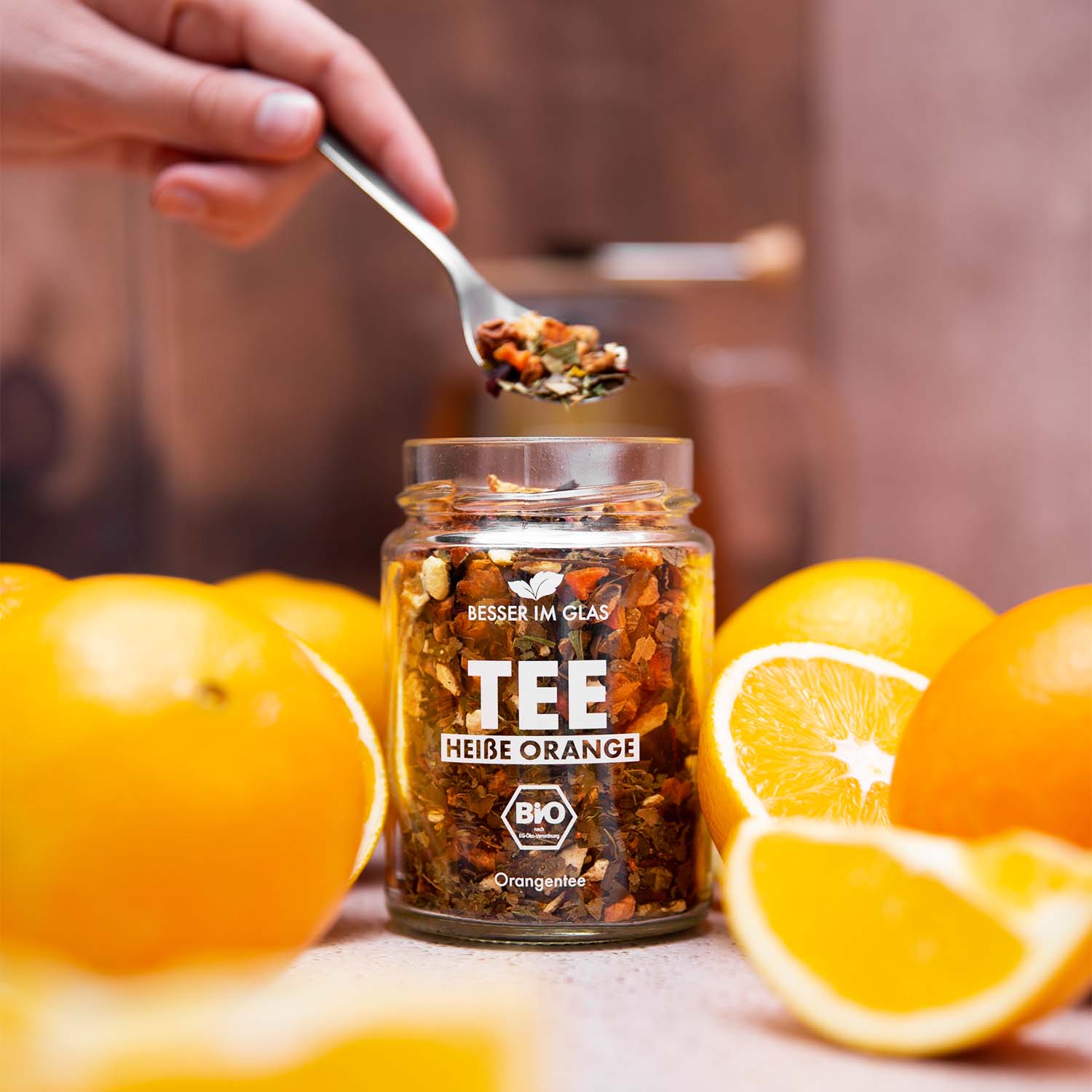 Loser bio Orangen Tee im Glas mit Orangen-Stücken. Bio-Früchte-Kräutertee mit Bio Karotte aromatisiert mit Orangen- und Mandarinengeschmack. Umgeben von Orangen. Der Bio Tee wird mit einem Löffel aus dem Tee Glas genommen. Bio Tee ohne Zucker.