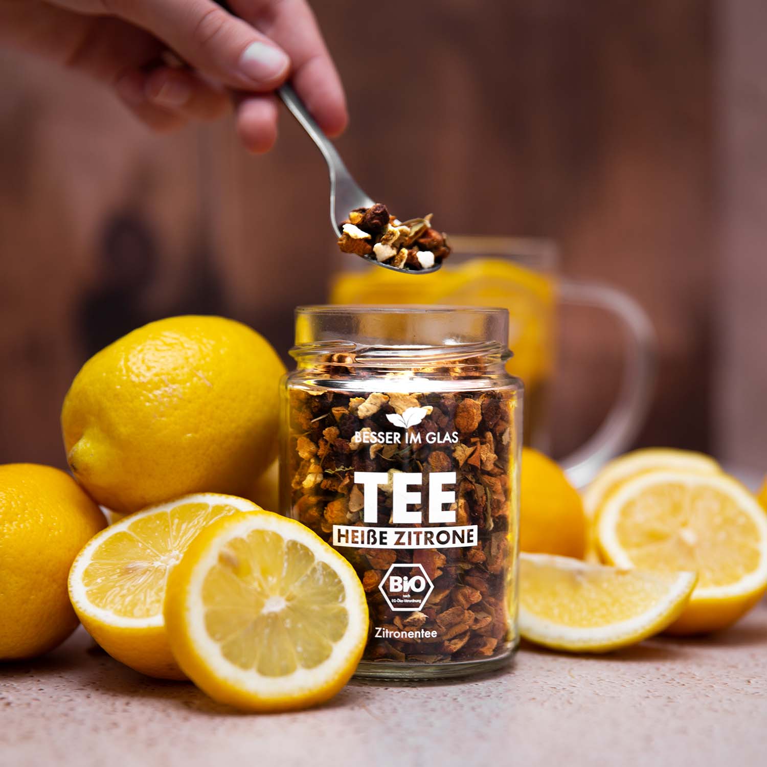 Loser bio Zitronen Tee im Glas mit Zitronen-Stücken. Bio-Früchte-Kräuterteemischung aromatisiert mit Zitronengeschmack. Umgeben von Zitronen. Der Bio Tee wird mit einem Löffel aus dem Tee Glas genommen. Außerdem ist eine Teetasse zu sehen. In dem Teeglas mit Henkel befinden sich Zitronenscheiben. Bio Tee ohne Zucker.