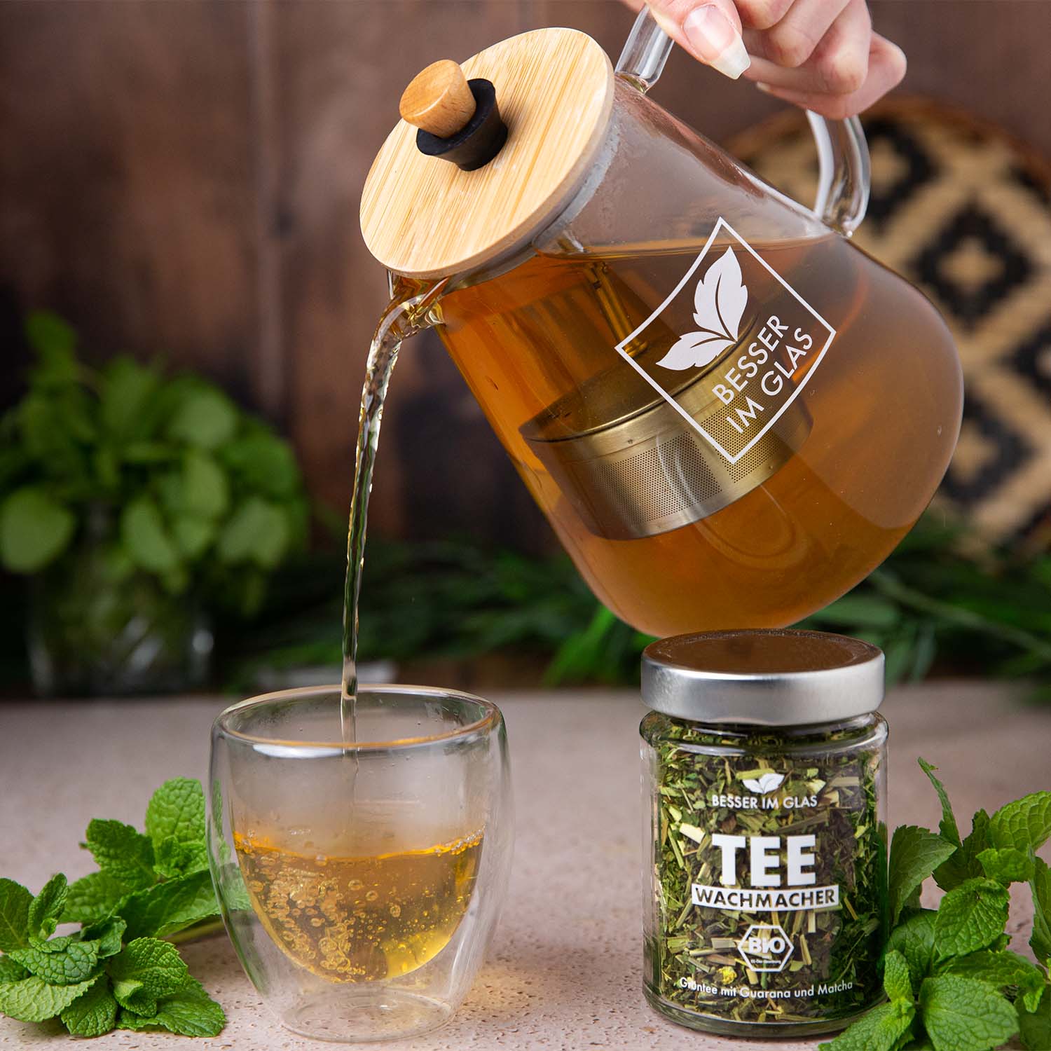 Loser bio Grün Tee im Glas. Bio-Mischung aus grünem Tee, Kräutern, Zitrusschalen, Guarana mit Limettengeschmack, natürlich aromatisiert. Das Tee Glas ist umgeben von Minze. Außerdem wird der gebrühte Wachmacher Grüntee aus einer Teekanne in ein leeres Glas geschüttet. In der Glaskanne befindet sich ein Teesieb mit abnehmbaren Bambus Deckel. Bio Tee ohne Zucker.