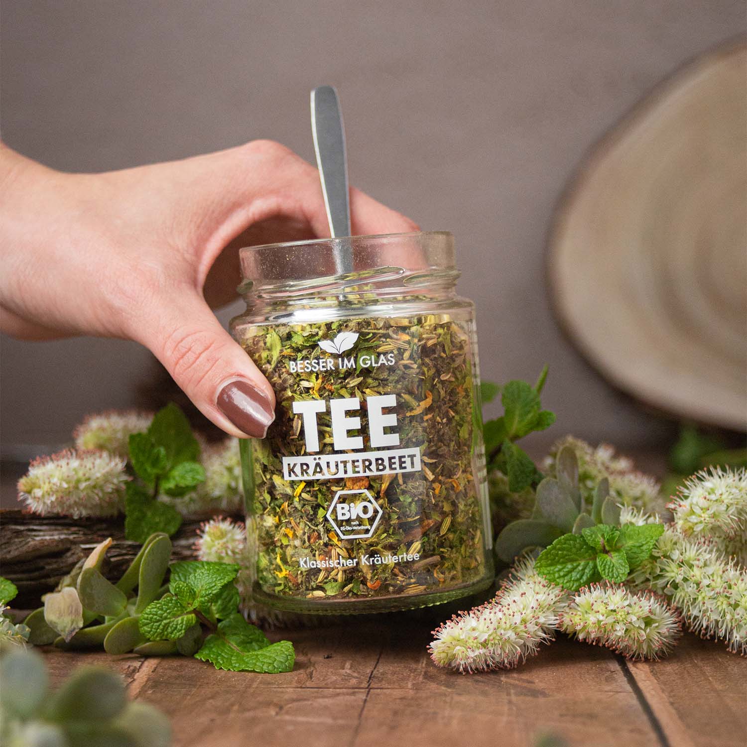 Loser bio Kräuter Tee im Glas mit Kräuter Stücken. Bio-Kräutertee mit Bio-Apfel. Das Tee Glas ist umgeben von Minze und Kräutern. Der Bio Tee wird mit einem Löffel aus dem Tee Glas genommen. Das Teeglas wird mit einer Hand hochgehoben. Bio Tee ohne Zucker.