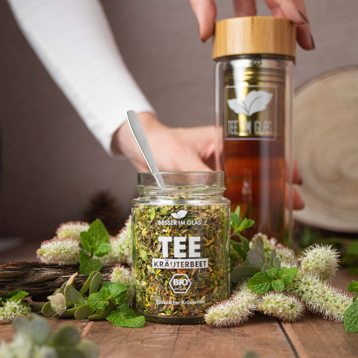 Loser bio Kräuter Tee im Glas mit Kräuter Stücken. Bio-Kräutertee mit Bio-Apfel. Das Tee Glas ist umgeben von Minze und Kräutern. Der Bio Tee wird mit einem Löffel aus dem Tee Glas genommen. Der gebrühte Tee befindet sich in einer To-Go Glasflasche mit Bambusdeckel und Teesieb. Die Doppelwandige Glasflasche wird mit einer Hand gehalten und mit einer anderen am Holzdeckel verschlossen. Bio Tee ohne Zucker.