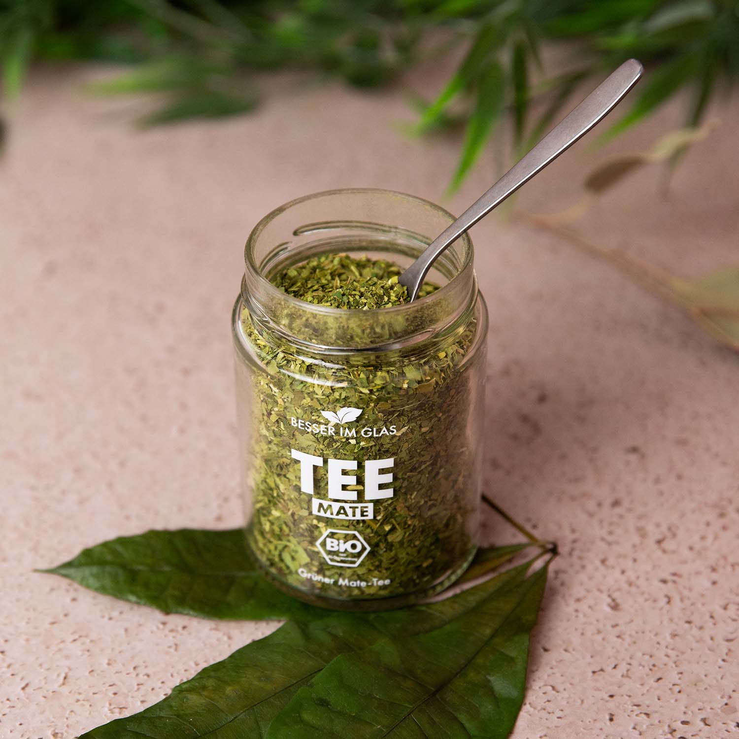 Loser bio Mate Tee im Glas. Bio-Matetee-Mischung. Der Bio Tee wird mit einem Löffel aus dem Tee Glas genommen. Das Tee Glas ist umgeben von Blättern. Bio Tee ohne Zucker.