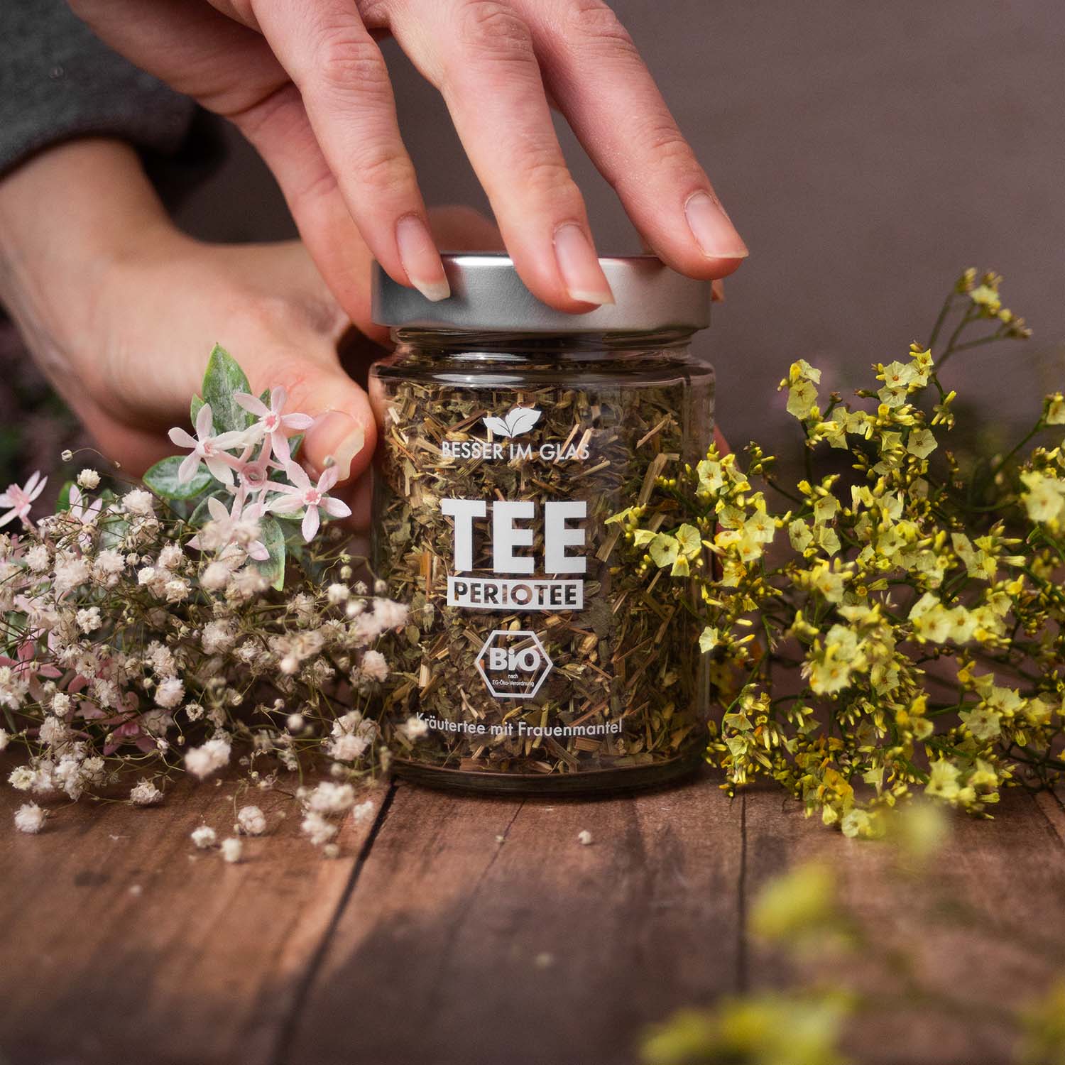 Loser bio Kräuter Tee im Glas. Bio-Kräutertee aromatisiert mit Grapefruit- und Limettengeschmack. Der Tee enthält Bio-Melisse, Bio-Zitronengras, Bio-Schafgarbenkraut, Bio-Rosmarin, Bio-Karotte, Bio-Kamille, Bio-Spirulina, Bio-Frauenmantel, Bio-Süßkraut, Bio-Ringelblumenblüten, Bio-Rotkleeblüten, natürliches Grapefruitaroma und Limettenöl. Das Tee Glas ist umgeben von Blumen. Der Bio Tee wird mit einem Löffel aus dem Tee Glas entnommen. Bio Tee ohne Zucker.