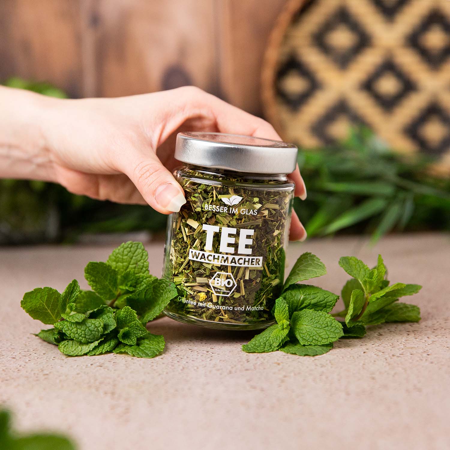 Loser bio Grün Tee im Glas. Bio-Mischung aus grünem Tee, Kräutern, Zitrusschalen, Guarana mit Limettengeschmack, natürlich aromatisiert. Das Tee Glas ist umgeben von Minze und wird von einer Hand in einem schrägen Winkel gehalten. Bio Tee ohne Zucker.