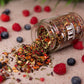 Loser bio Früchtetee im Glas mit Apfel und Früchte Stücken. Bio-Früchtetee-Mischung mit Himbeer- und Erdbeergeschmack, natürlich aromatisiert. Das Tee Glas liegt auf dem Tisch und ist umgeben von Himbeeren und Blaubeeren. Ein Teil des Früchtetees ist ausgeschüttet. Bio Tee ohne Zucker.