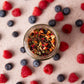 Loser bio Früchtetee im Glas mit Apfel und Früchte Stücken. Bio-Früchtetee-Mischung mit Himbeer- und Erdbeergeschmack, natürlich aromatisiert. Das Tee Glas ist umgeben von Himbeeren und Blaubeeren. Bio Tee ohne Zucker.